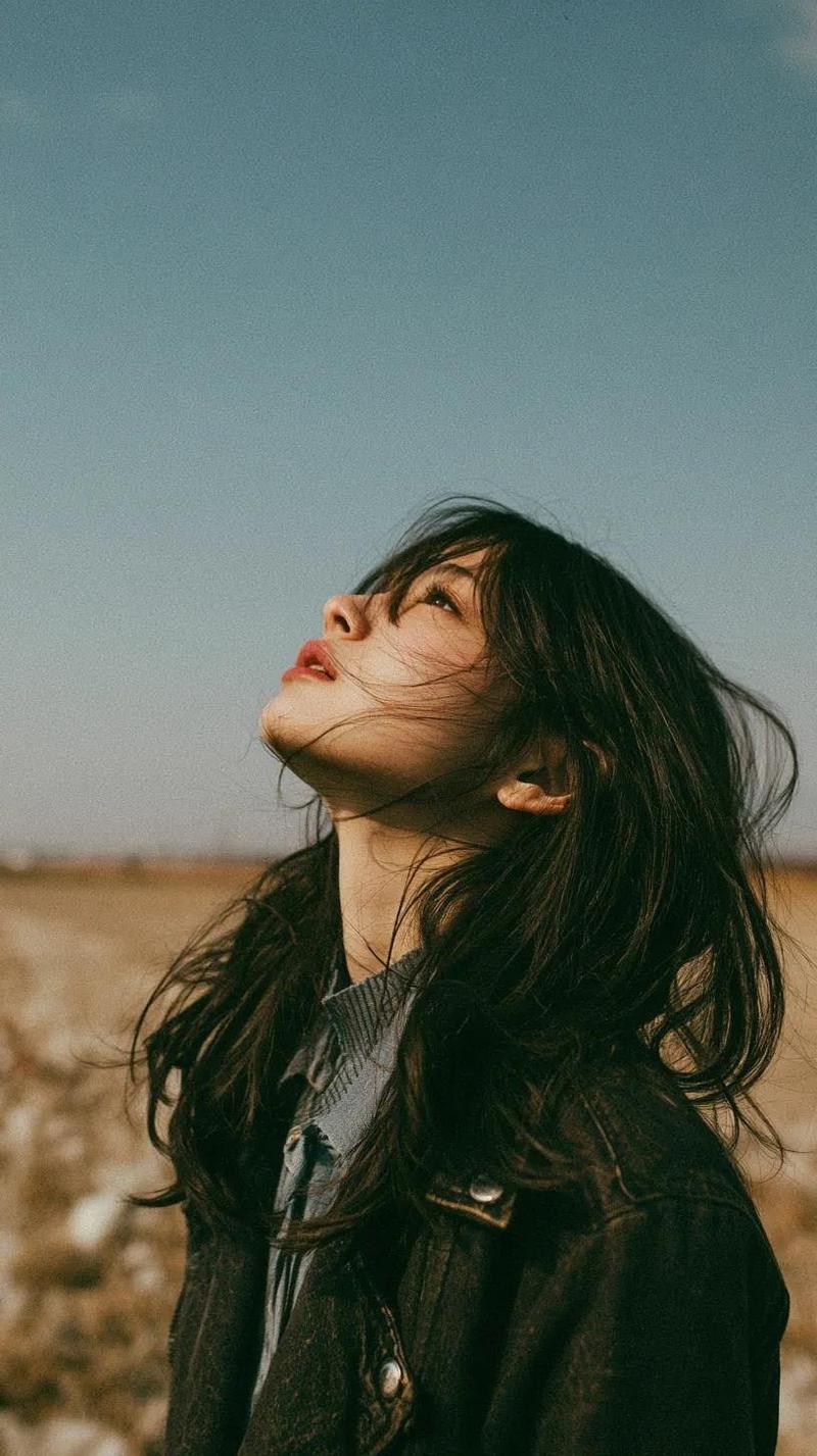 一位美丽的年轻东亚女性独自站在开阔的野花草甸中,头顶是无尽的蓝天,她闭眼或半睁眼向上凝视,仿佛在拥抱微风。长长的自然凌乱黑发自由地披散在脸上,柔...